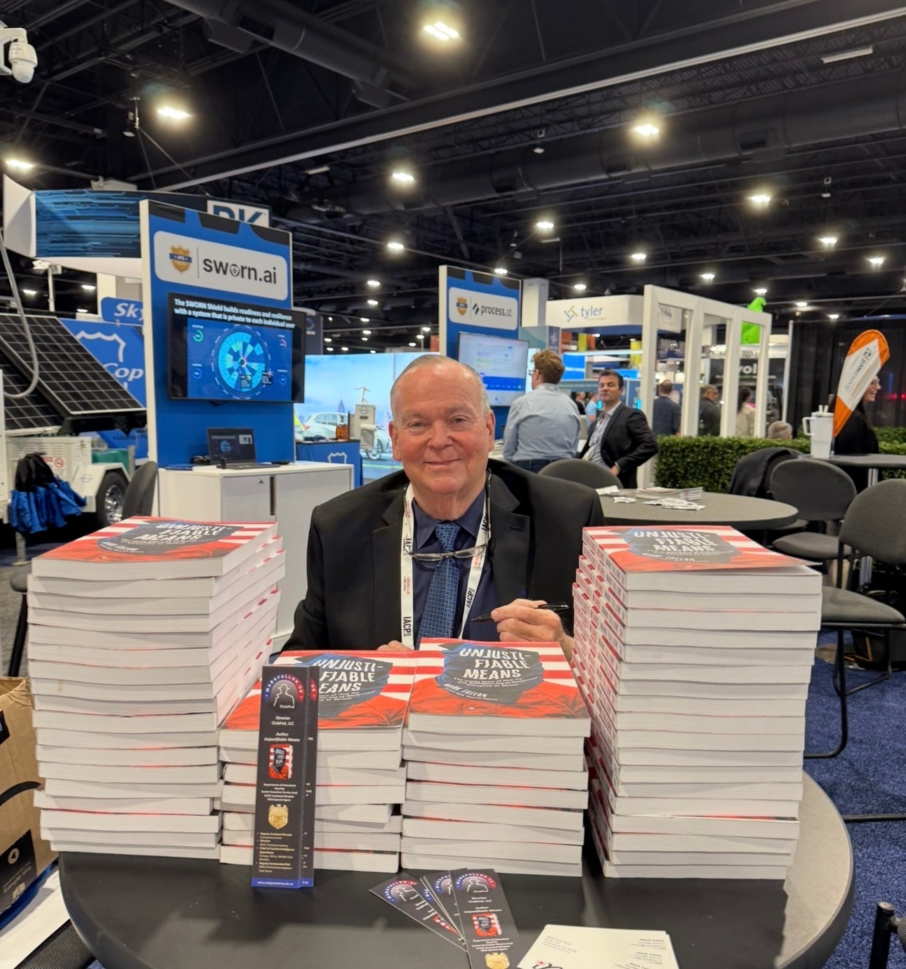 Mark Fallon at a book signing booth at a law enforcement conference, surrounded by attendees holding copies of Unjustifiable Means