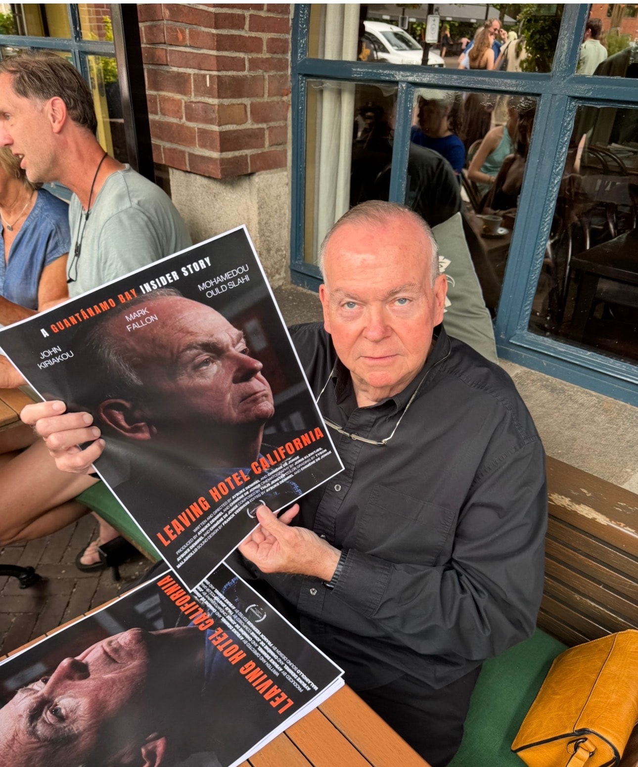 Mark Fallon holding a Leaving Hotel California film poster at an outdoor café