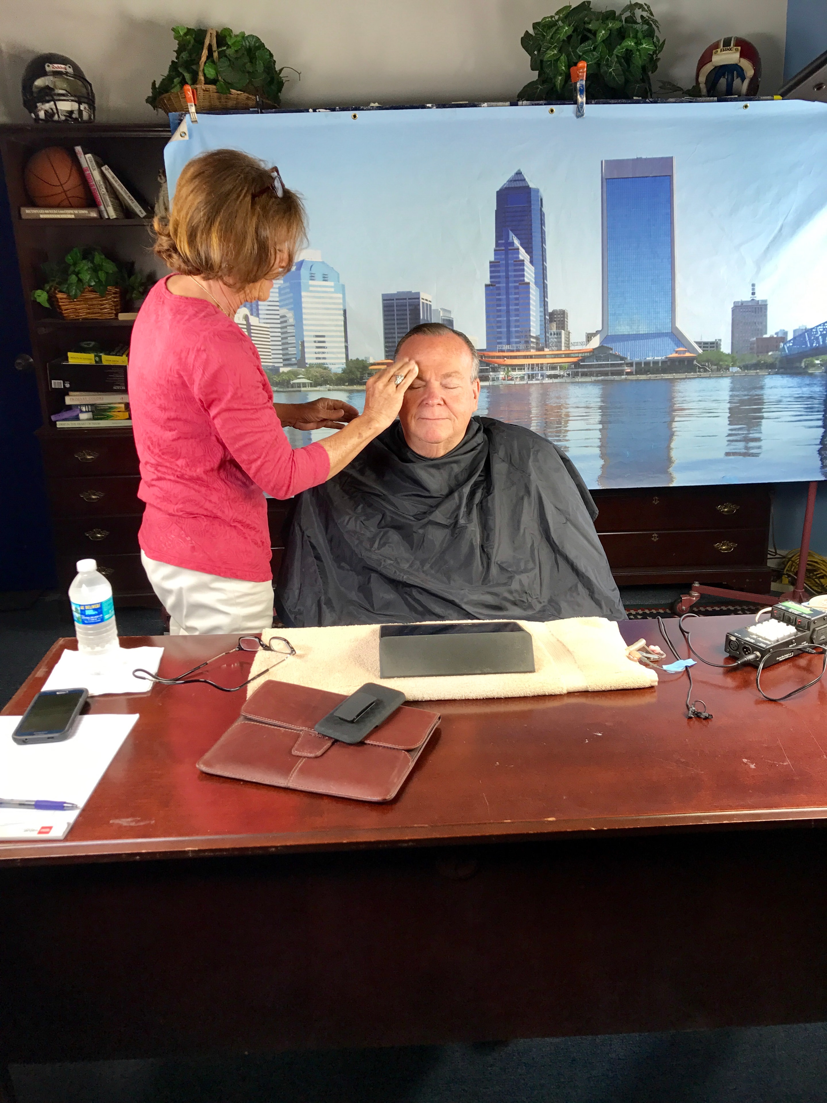 Mark Fallon getting makeup applied before a TV shoot, seated at a desk with a Jacksonville skyline backdrop
